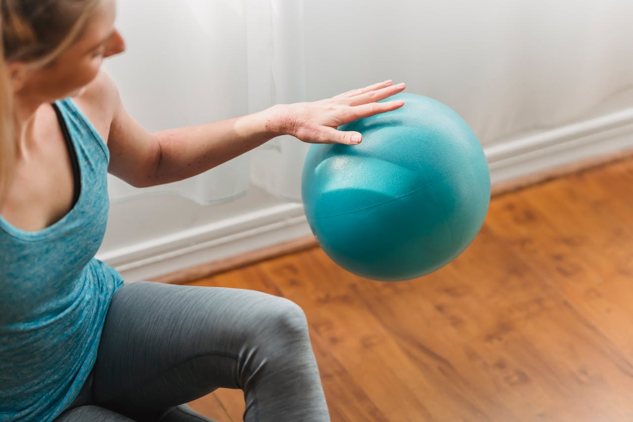 From above of crop young slim woman in activewear sitting on laminate of modern fitness studio and hitting gymnastic ball on floor